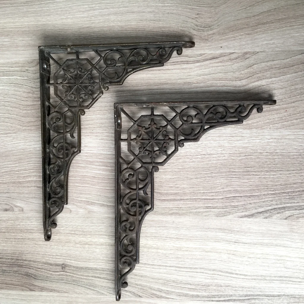 Two ornate dark metal scrollwork brackets with geometric medallions resting on a light wood-grain surface, conveying craftsmanship and variety